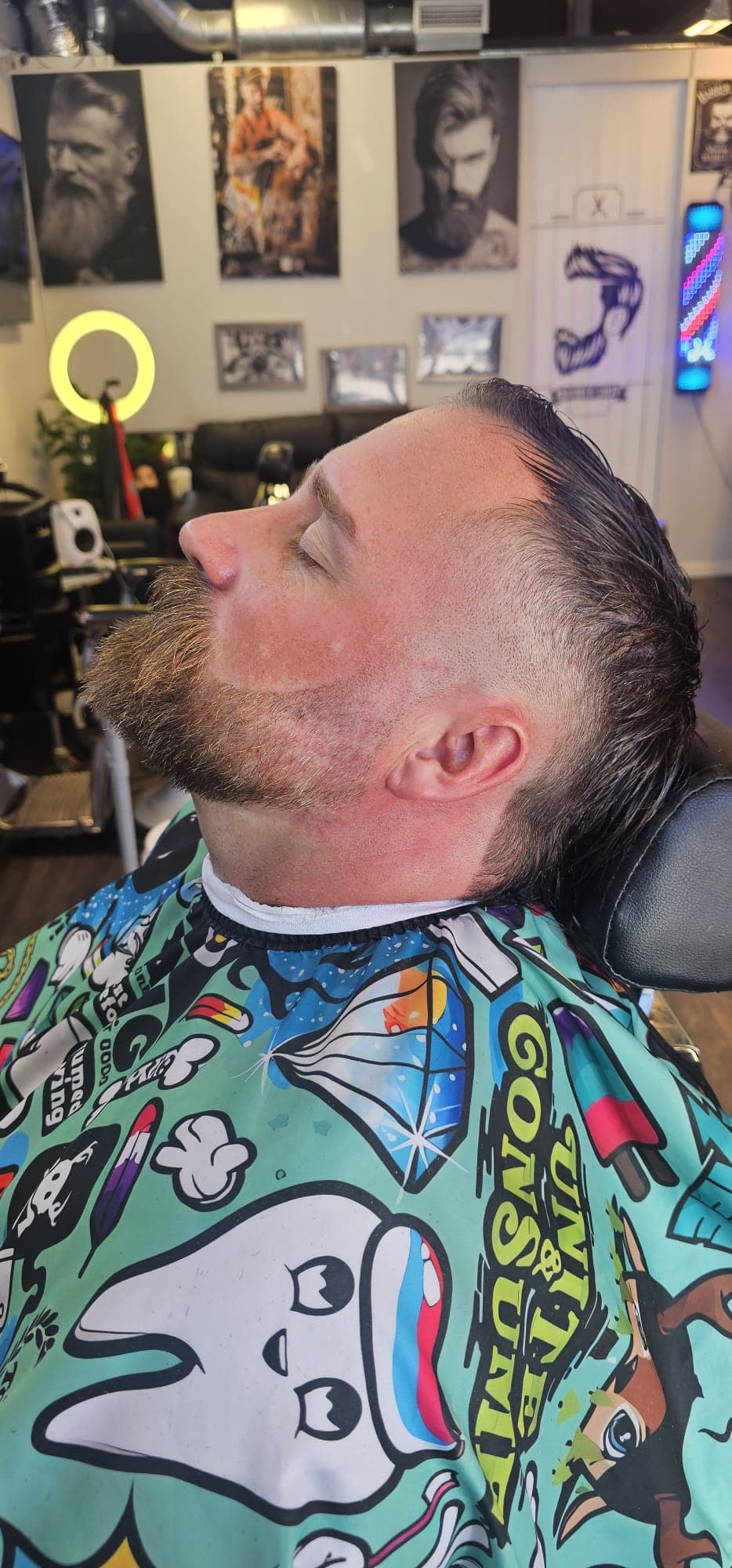 Trimmat skägg och bakåtkammat hår med fade, i profil framför salongens vägg med affischer och barberarstolpe